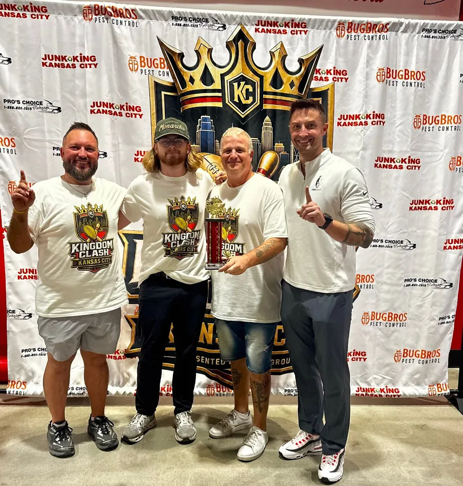 Four men in Kingdom Clash shirts pose with a red and gold trophy in front of the Kingdom Clash Kansas City backdrop, two of them pointing a finger up